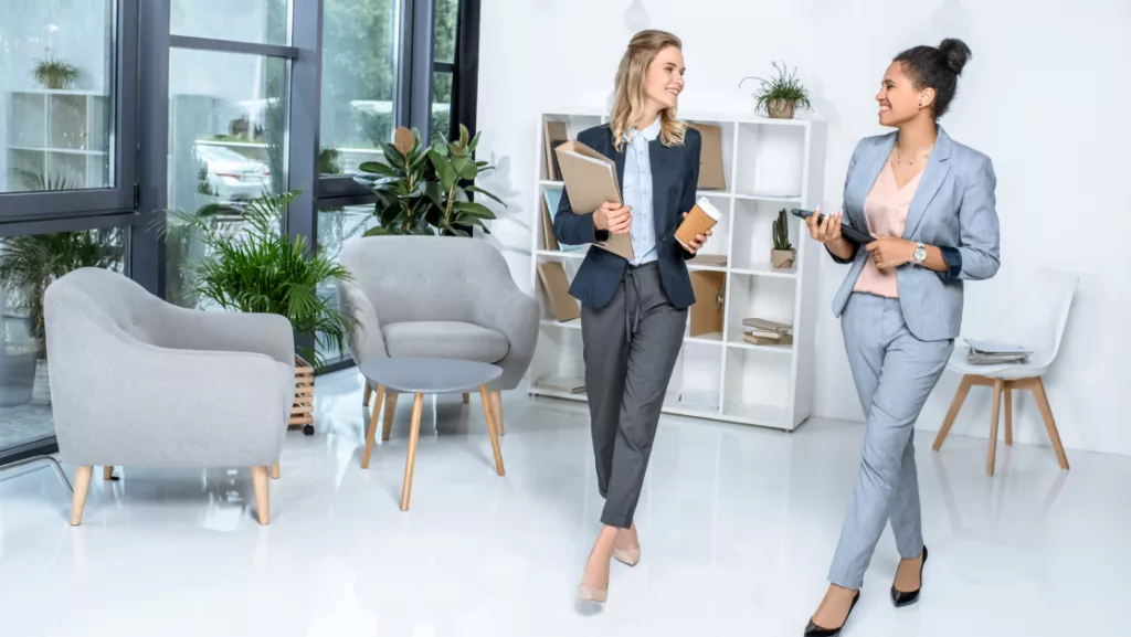 Two professional women in business attire walking and talking in a modern office lobby.